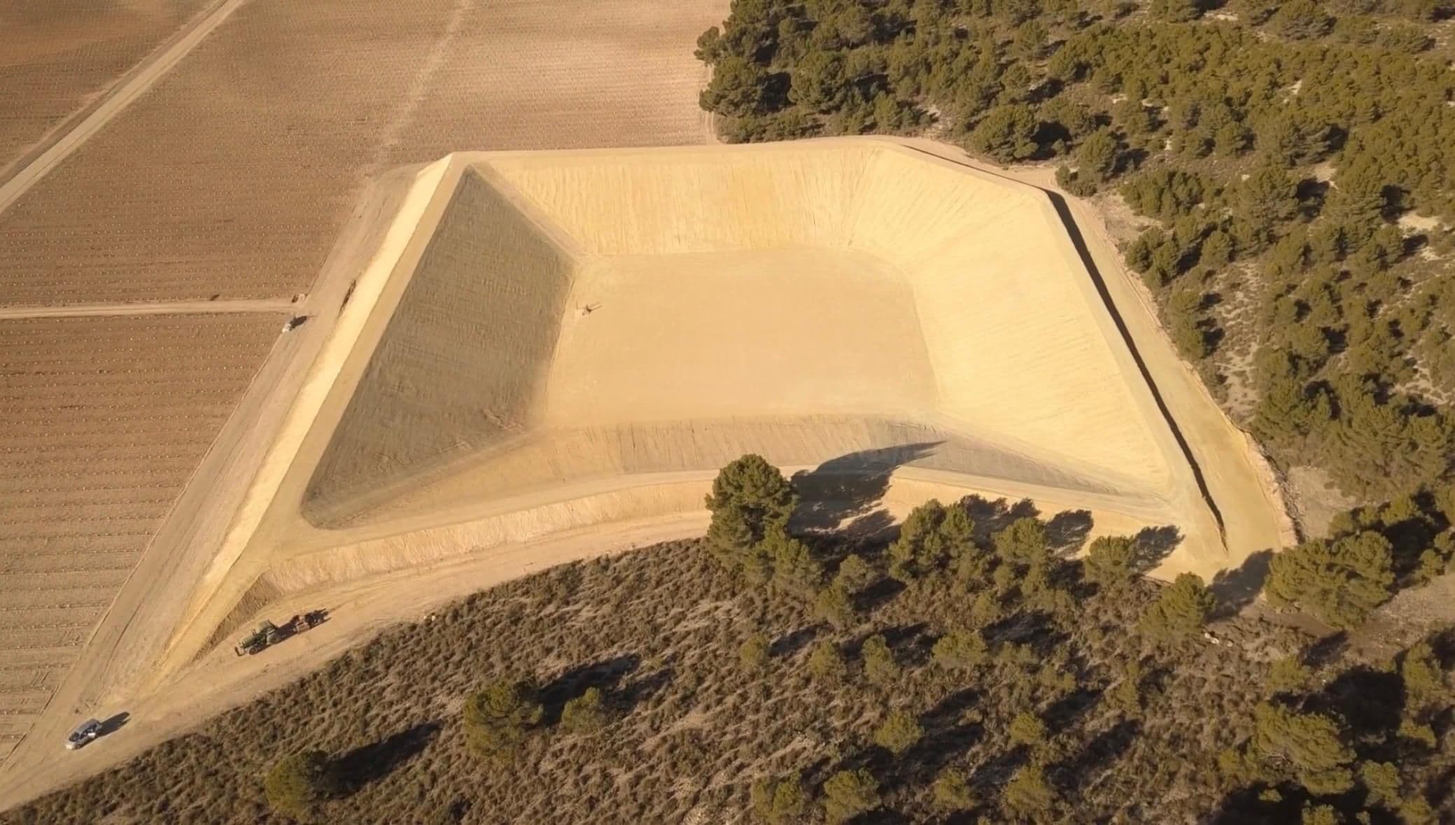 Plano amplio de la balsa terminada, con la viña a un lado y el pinar al otro