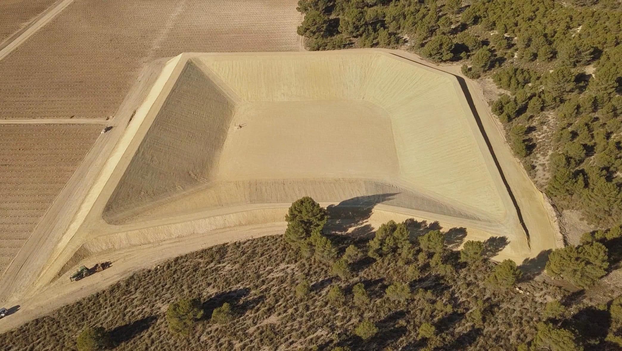Vista cenital de la balsa terminada, con los cuatro taludes perfilados en abanico, la pista de coronación alrededor y un tractor parado en el camino