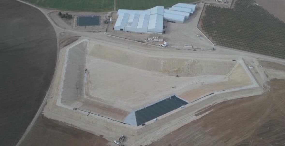 Vista aérea del vaso de la balsa ya excavado y perfilado, con la pista de coronación cerrada alrededor, los primeros paños de lámina impermeable extendidos en el fondo y las naves de la finca detrás