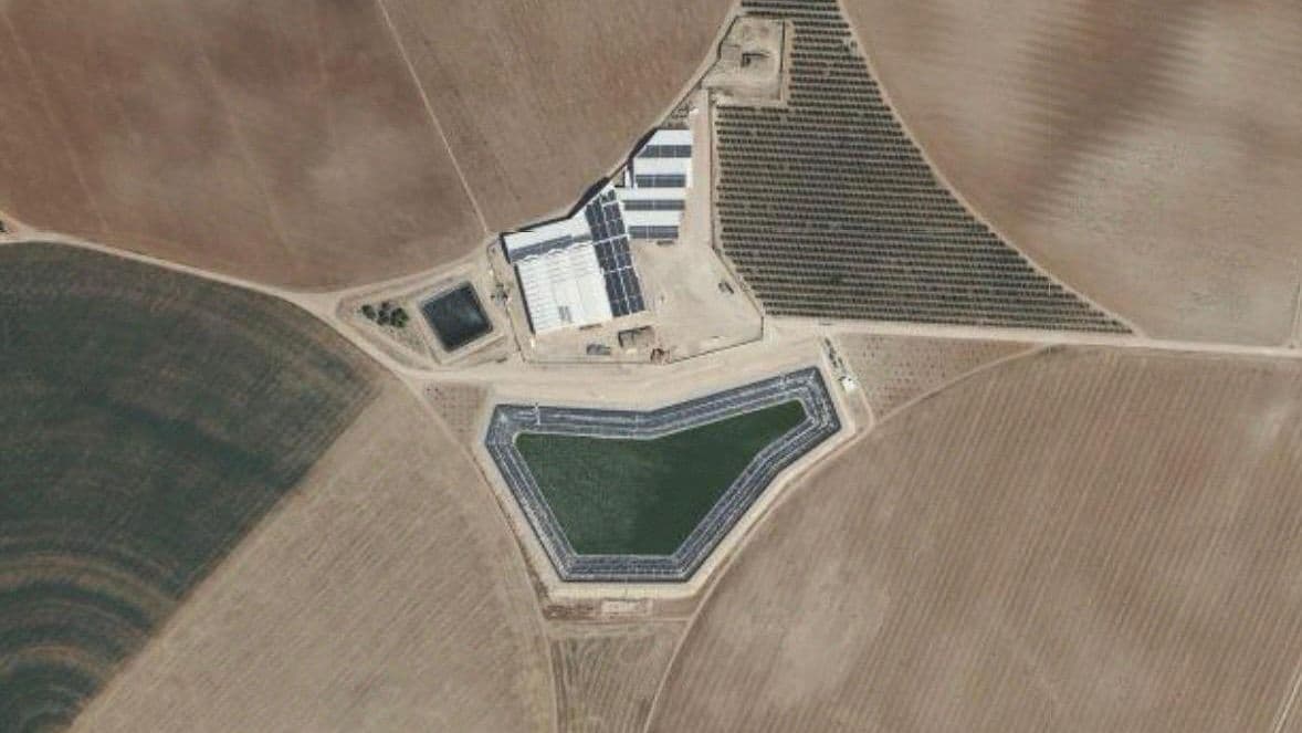 Vista cenital de la balsa terminada junto a las naves de la finca con placas solares, entre los campos de labor y una plantación de leñosos